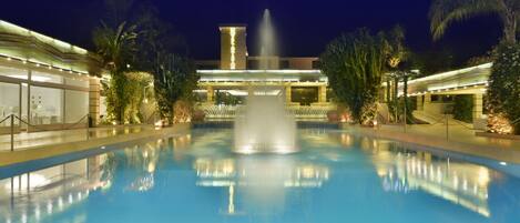Ein Poolbereich bei Nacht mit einer zentralen Fontäne, umgeben von Palmen und einem Gebäude mit beleuchteten Fenstern.
