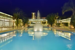 Zona de piscina nocturna con una fuente central, rodeada de palmeras y un edificio con ventanas iluminadas.