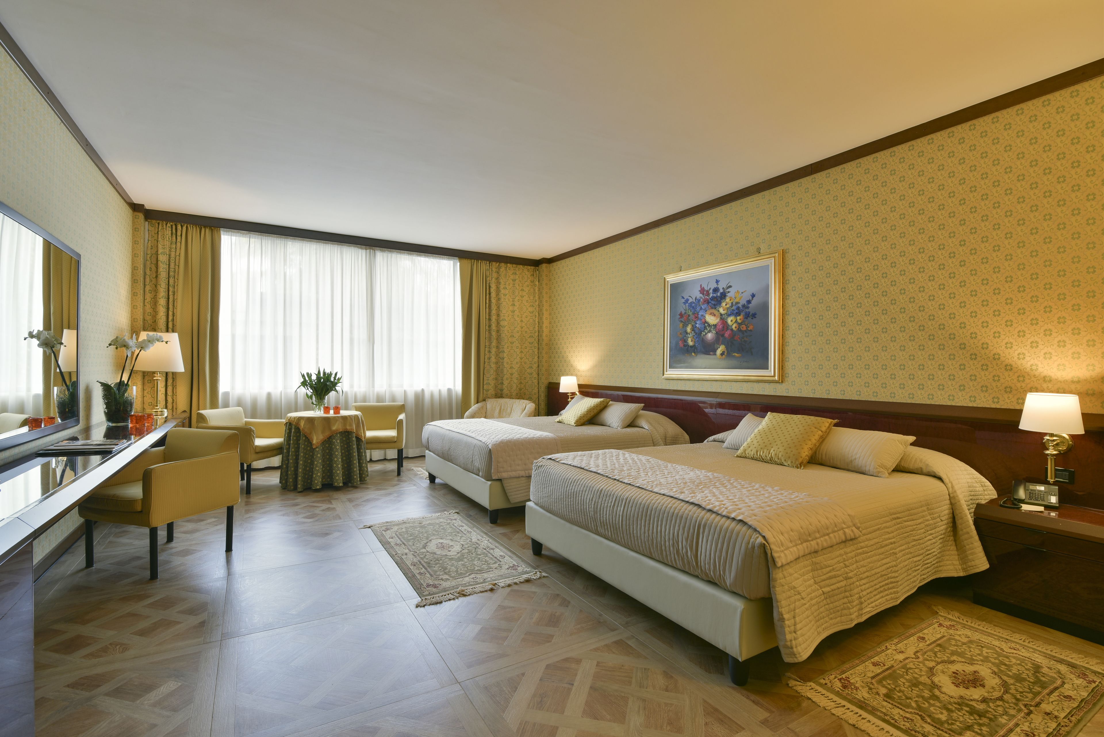A hotel room with two beds, a sitting area with a chair and a table, a desk with a lamp, and a large window with curtains.