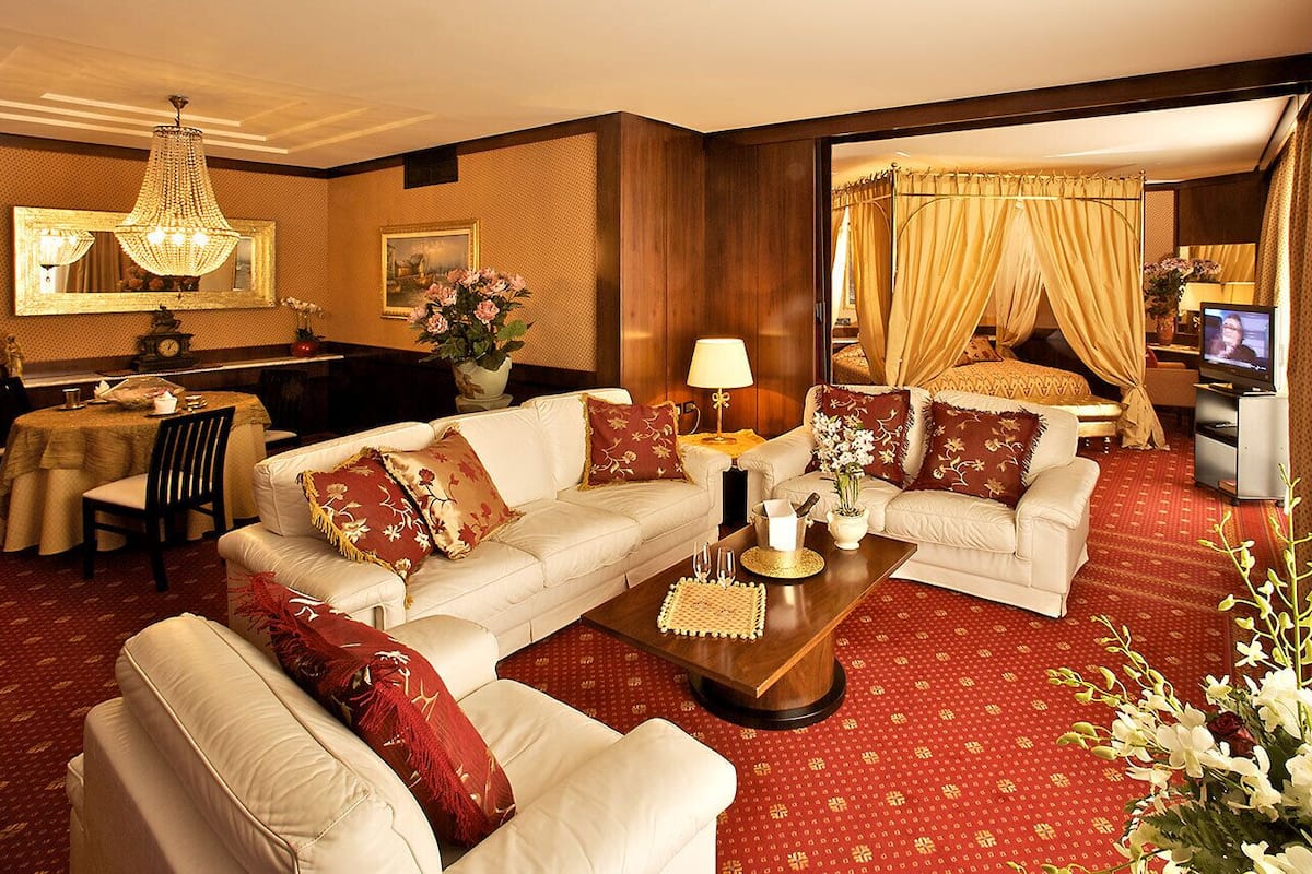 A hotel room with a large sofa set, a dining area with a table and chairs, a television, and a large mirror.