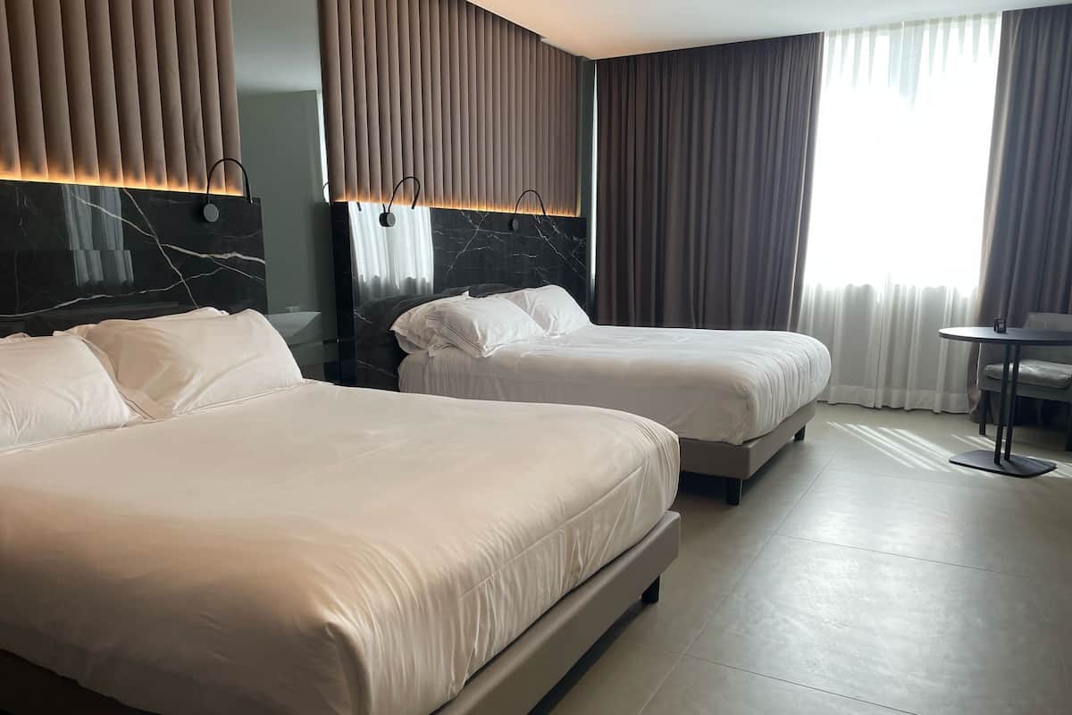 A hotel room with two beds, a marble headboard, a small round table, and a view of the outdoors.