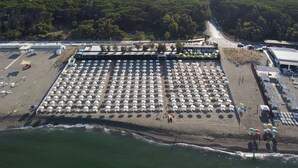 Vista aérea de una playa con hileras de sombrillas y sillas de playa blancas, un aparcamiento y vistas de la ciudad y las colinas al fondo.