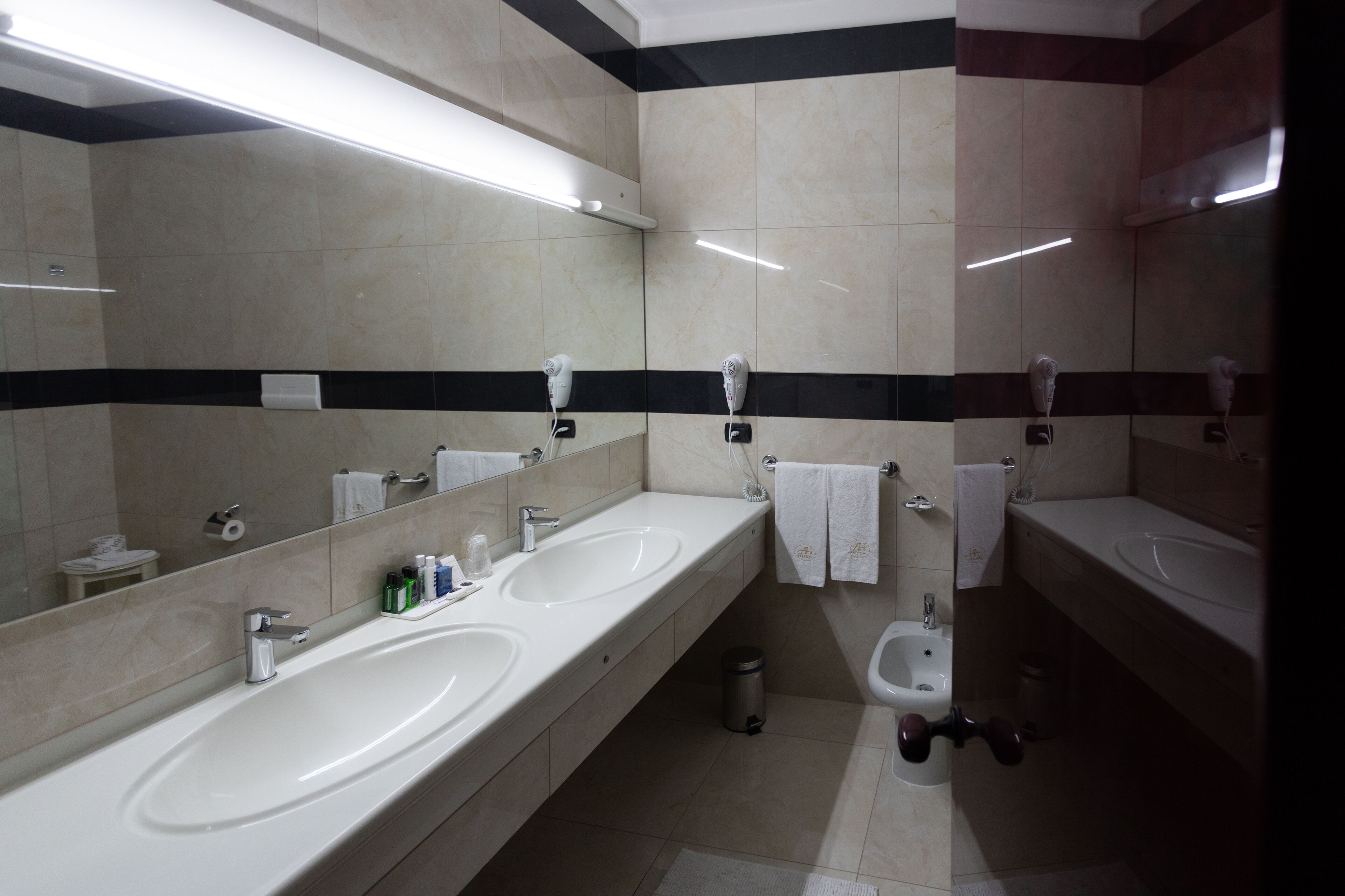 A modern bathroom with two sinks, a toilet, and a bidet. It features a large mirror, black and white striped walls, and a trash bin.
