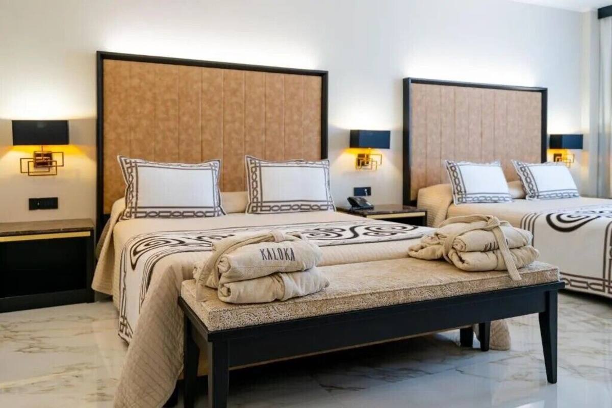 A hotel room with two beds, each with a headboard, bedside lamps, and a bedside table. There is a large mirror on the wall and a marble floor.