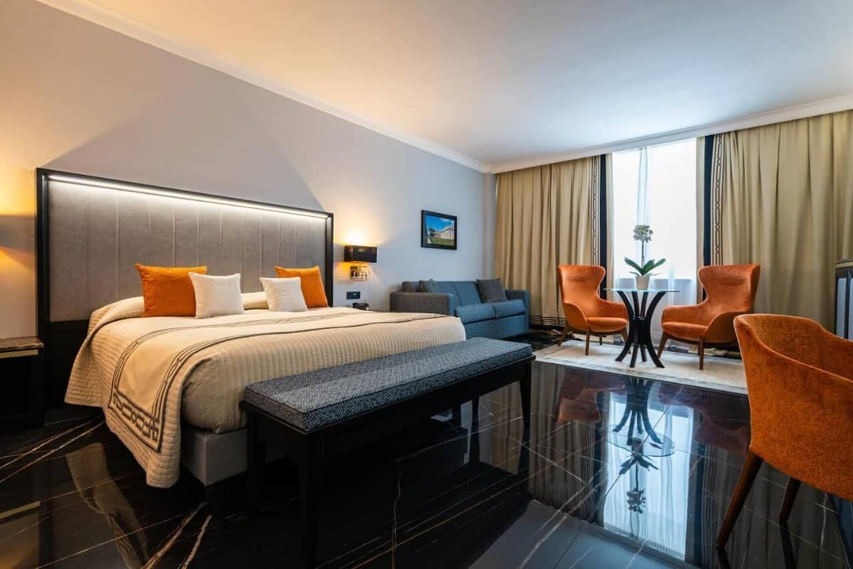 A hotel room with a large bed, a sofa, two armchairs, a small table, and a bench.