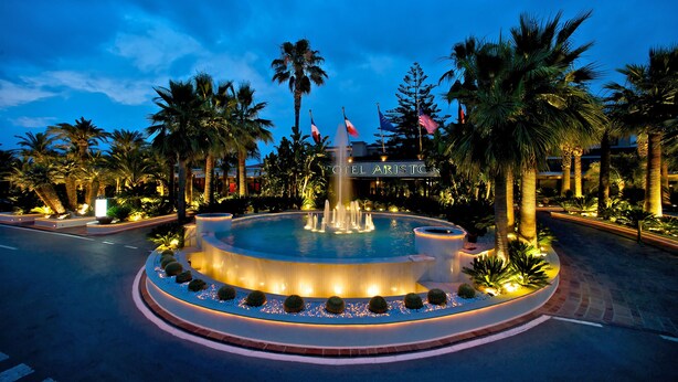 Una fuente circular con chorros de agua, rodeada de palmeras e iluminada por luces.