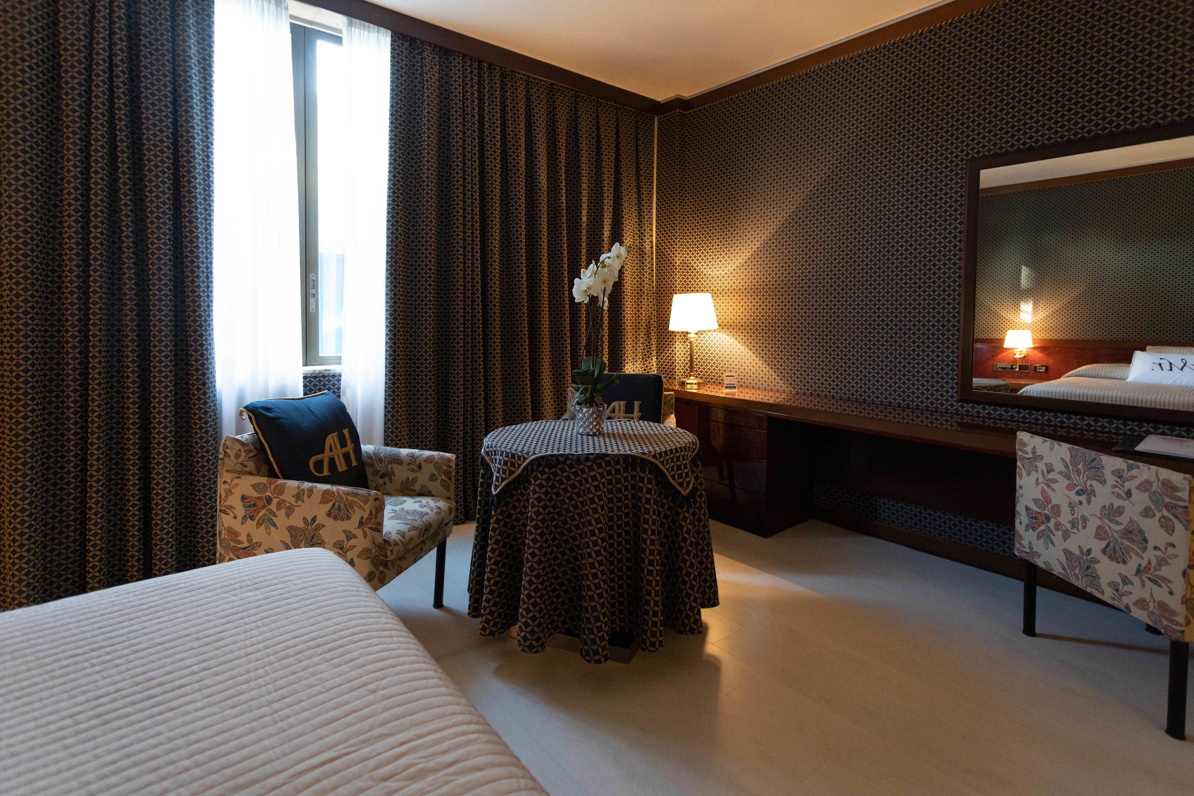 A hotel room with a bed, a desk with a lamp, a chair, and a small table.