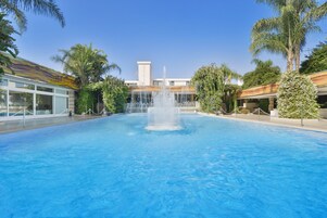 A swimming pool with a central fountain, surrounded by palm trees and a building with a modern design.