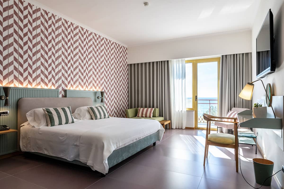 Ein modernes Hotelzimmer mit einem großen Bett, einem Schreibtisch mit Stuhl, einem Fernseher und Blick ins Freie.