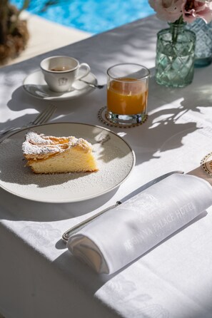 Una porción de pastel en un plato con azúcar glasé, un vaso de zumo de naranja y una taza de café sobre una mesa con vista a la piscina.