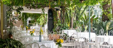 An outdoor dining area with white metal chairs and tables, surrounded by greenery and plants.