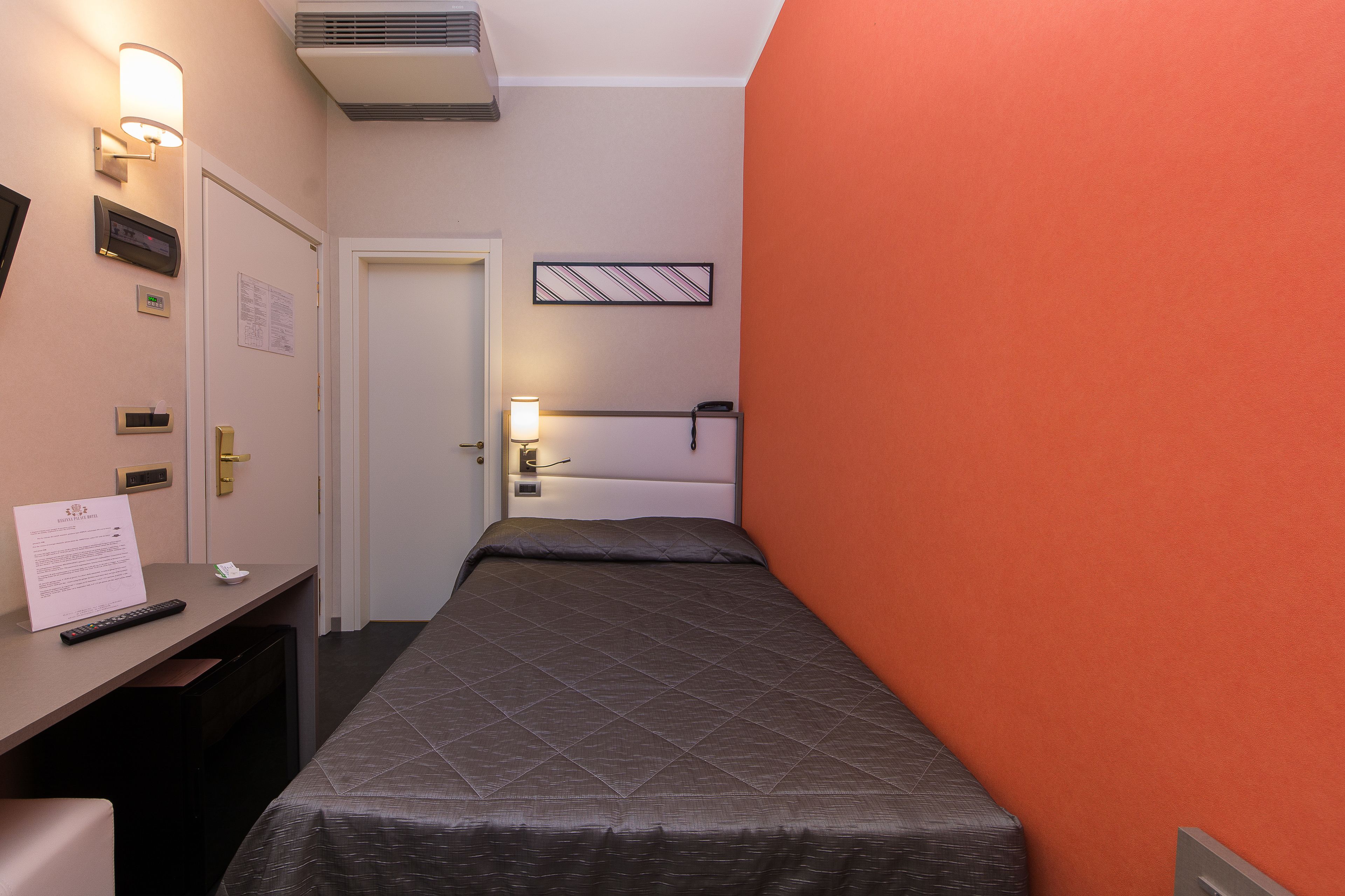 A hotel room with a bed, a desk with a remote control, a lamp, and a wall-mounted air conditioner.