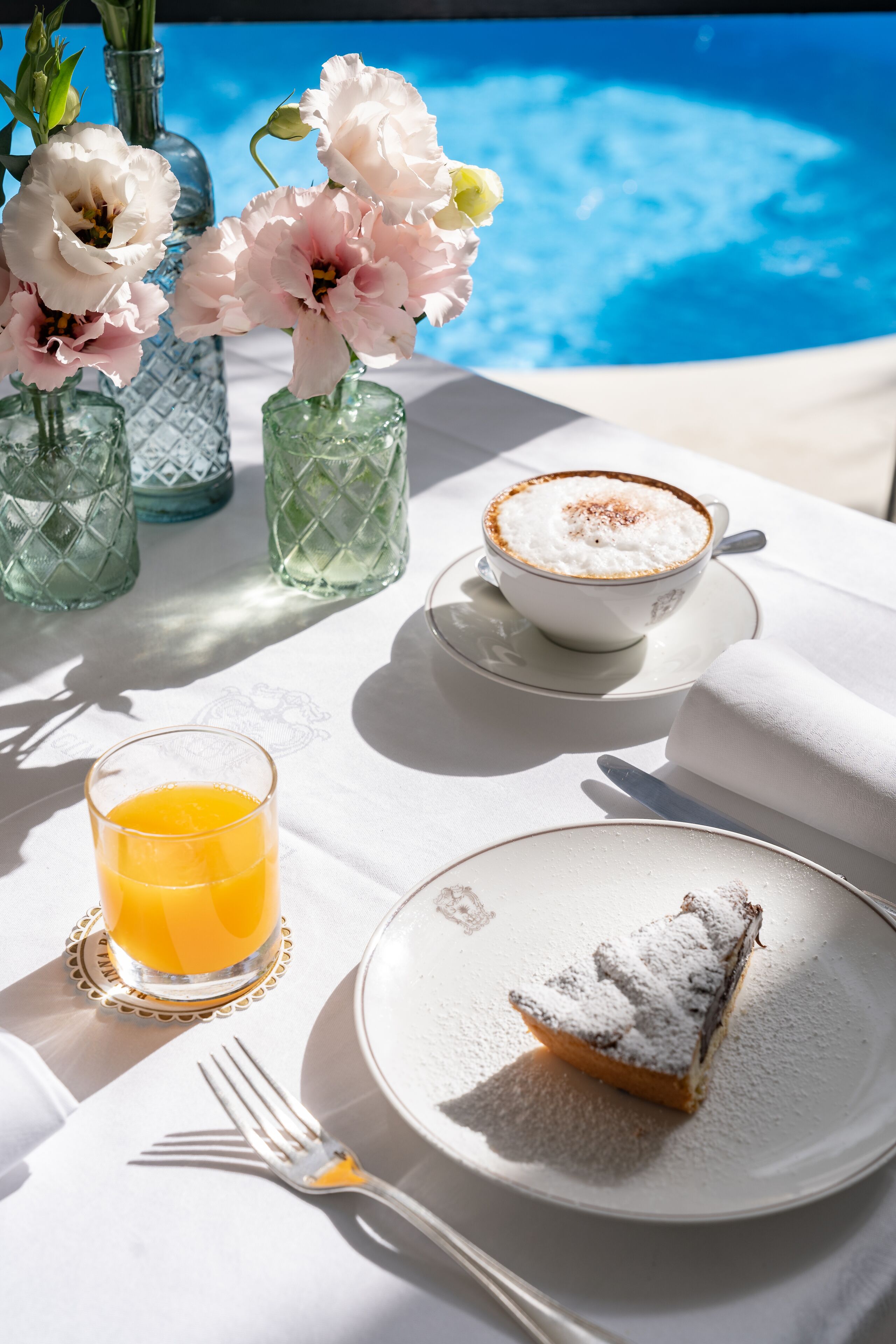 A table with a slice of cake, a glass of orange juice, a cup of coffee, and vases of flowers.
