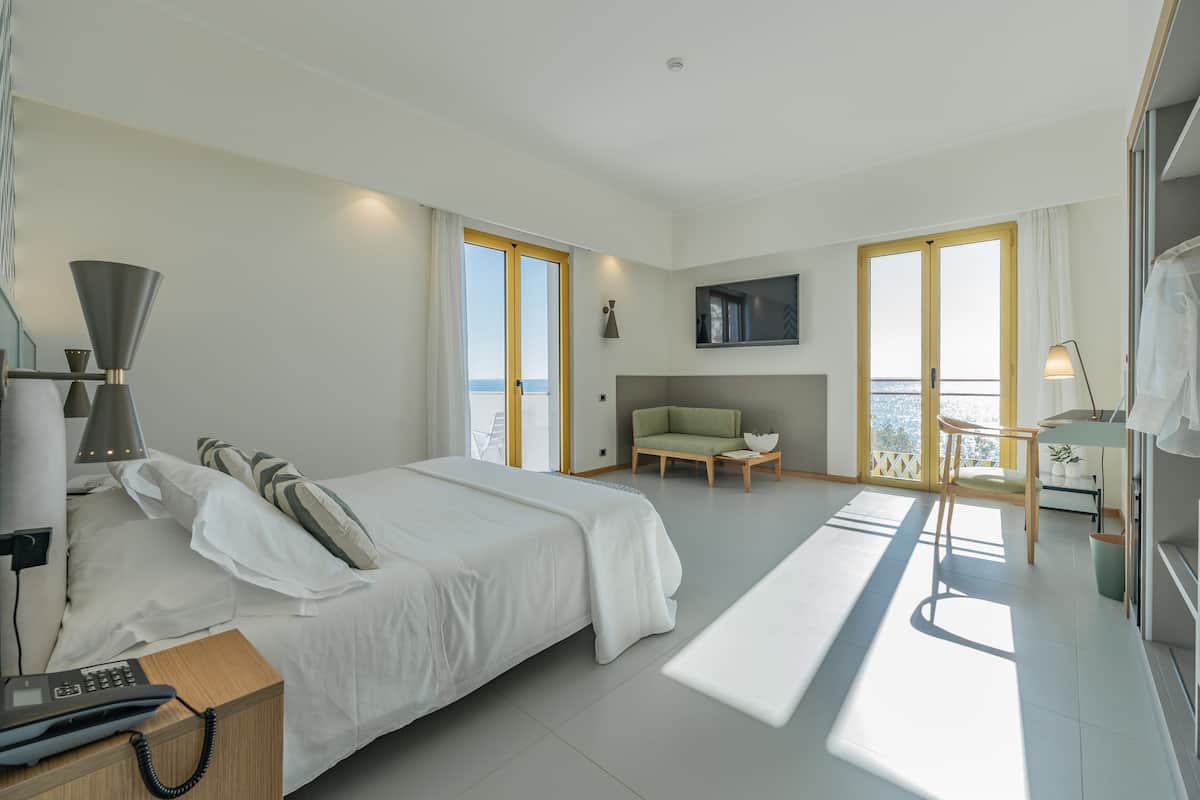 Ein modernes Hotelzimmer mit einem großen Bett, einem Schreibtisch mit Telefon, einem Sessel, einem Fernseher und einem Balkon mit Meerblick.