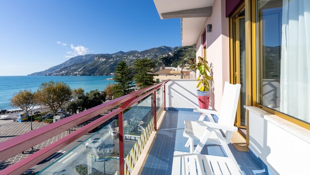 Un balcón con vistas despejadas al mar y las montañas, sillas blancas y una planta en maceta.