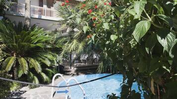 Zona de piscina con una barrera de cuerda, rodeada de abundante vegetación y un edificio al fondo.