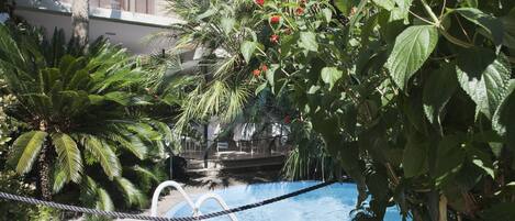Zona de piscina con una barrera de cuerda, rodeada de abundante vegetación y un edificio al fondo.