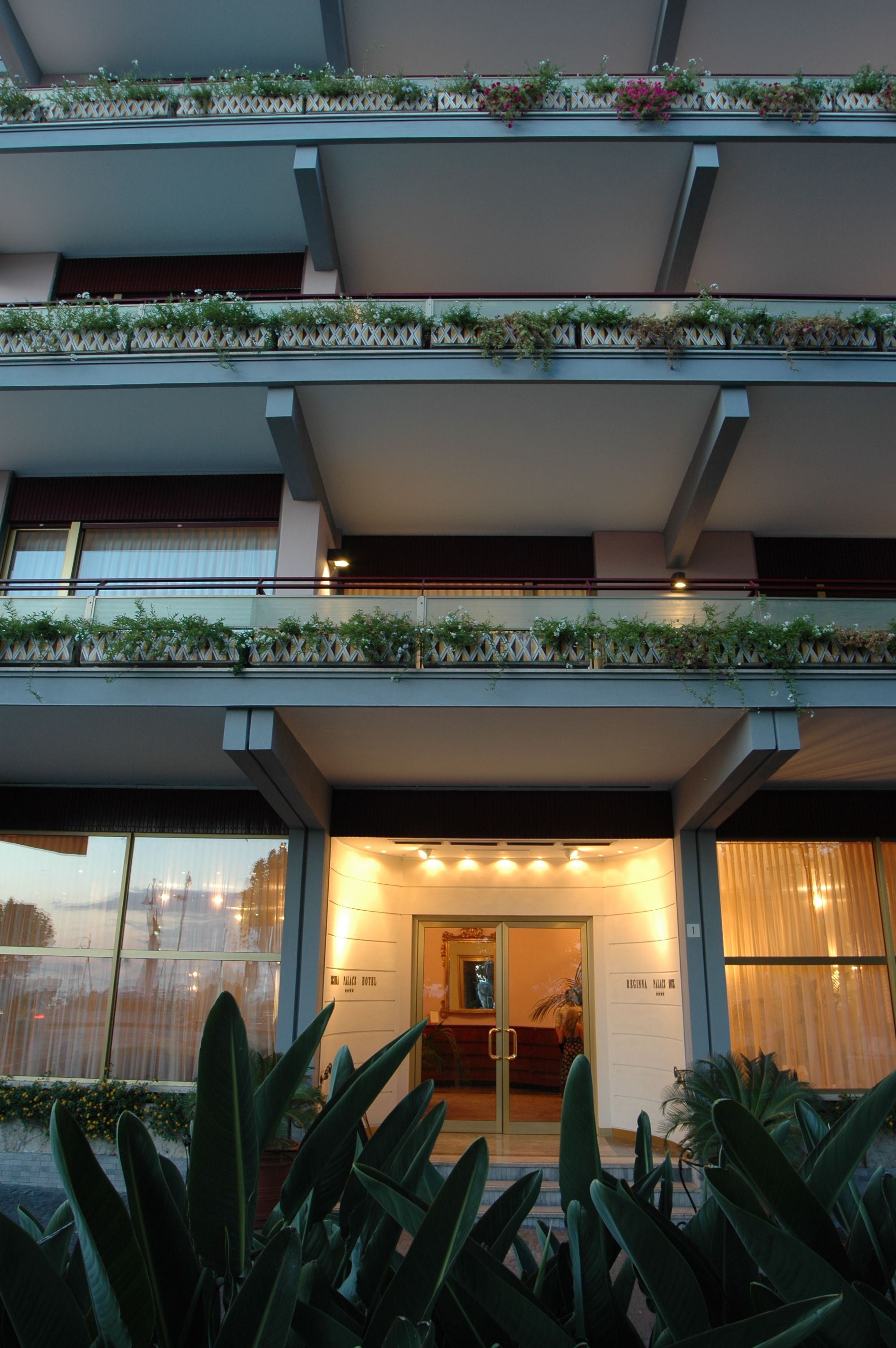 Een hotel met meerdere balkons, versierd met planten, grote ramen en een goed verlichte entree.