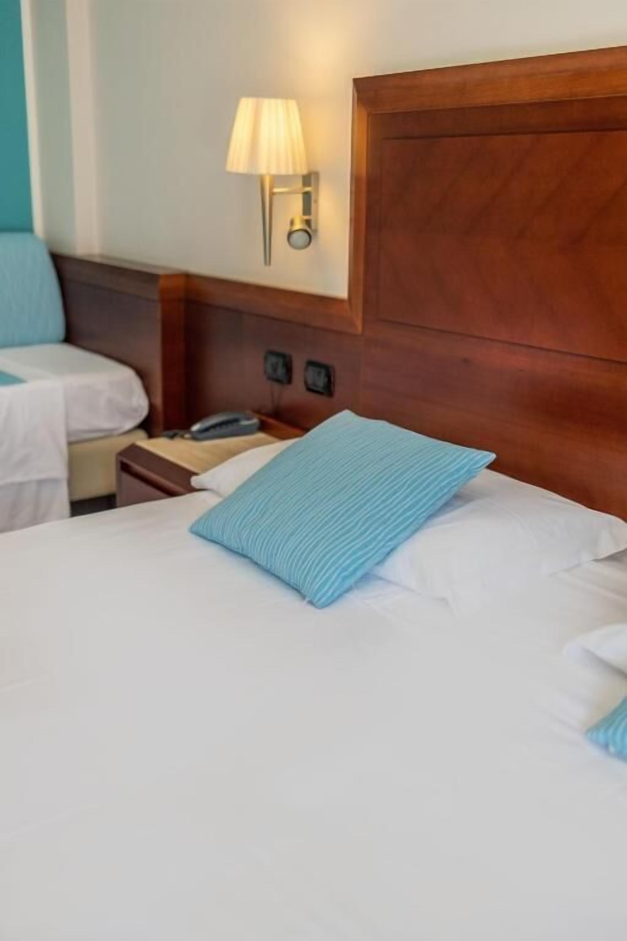 Habitación de hotel con cabecera de madera, lámpara de noche, teléfono y almohada azul sobre la cama.