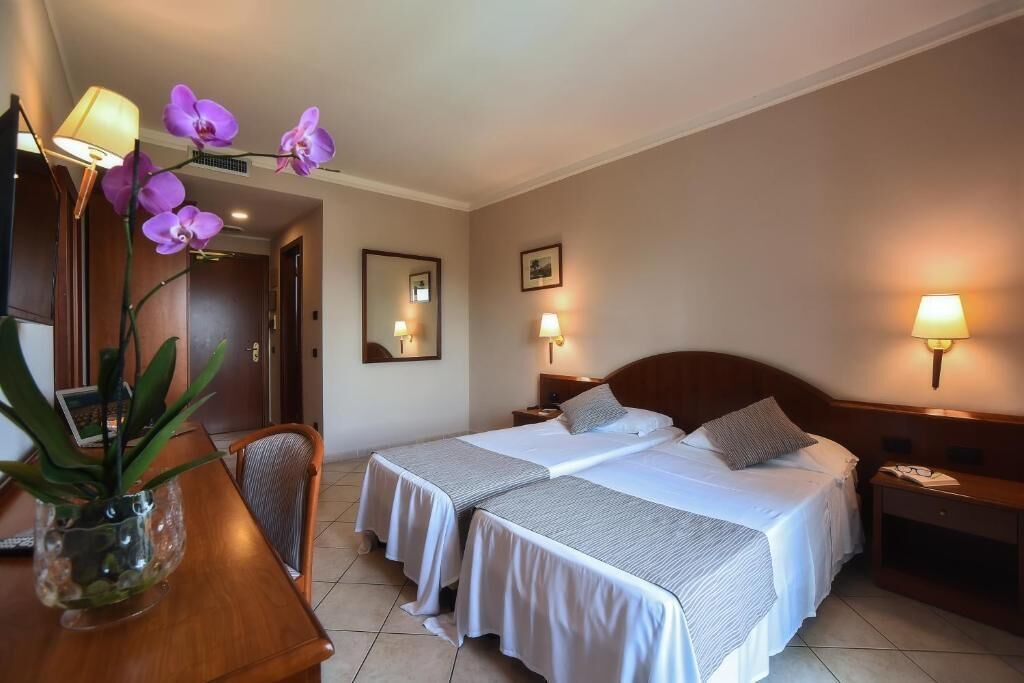 Habitación de hotel con dos camas, un escritorio con silla, un jarrón con flores y lámparas de pared.