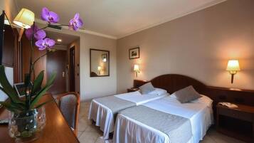 Habitación de hotel con dos camas, un escritorio con silla, un jarrón con flores y lámparas de pared.