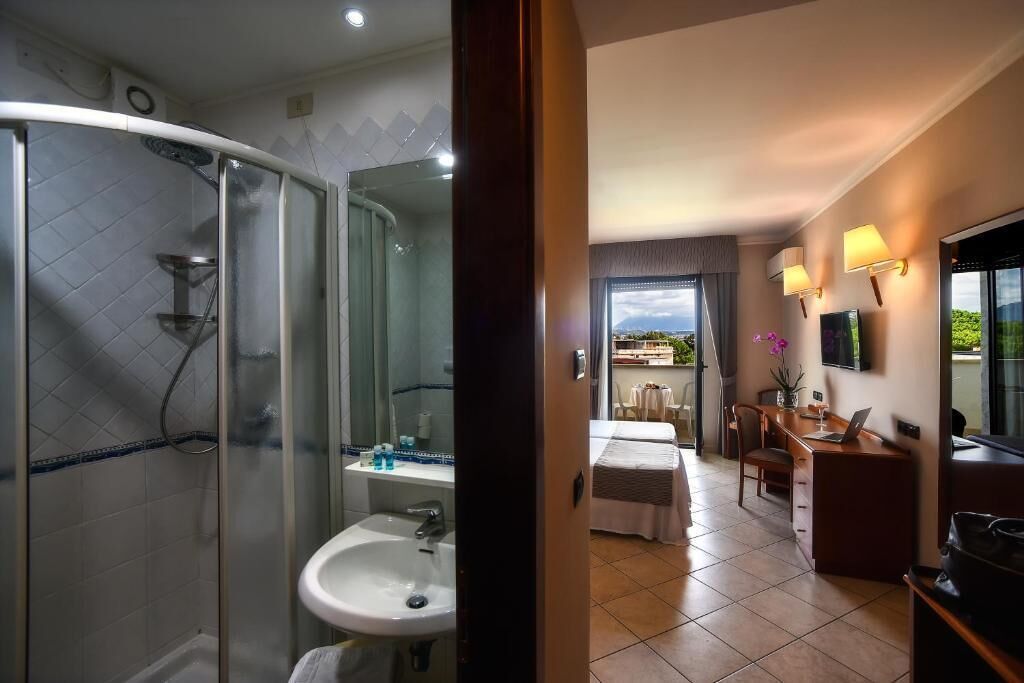 Habitación de hotel con ducha de vidrio, lavabo, cama, escritorio con silla y televisor.