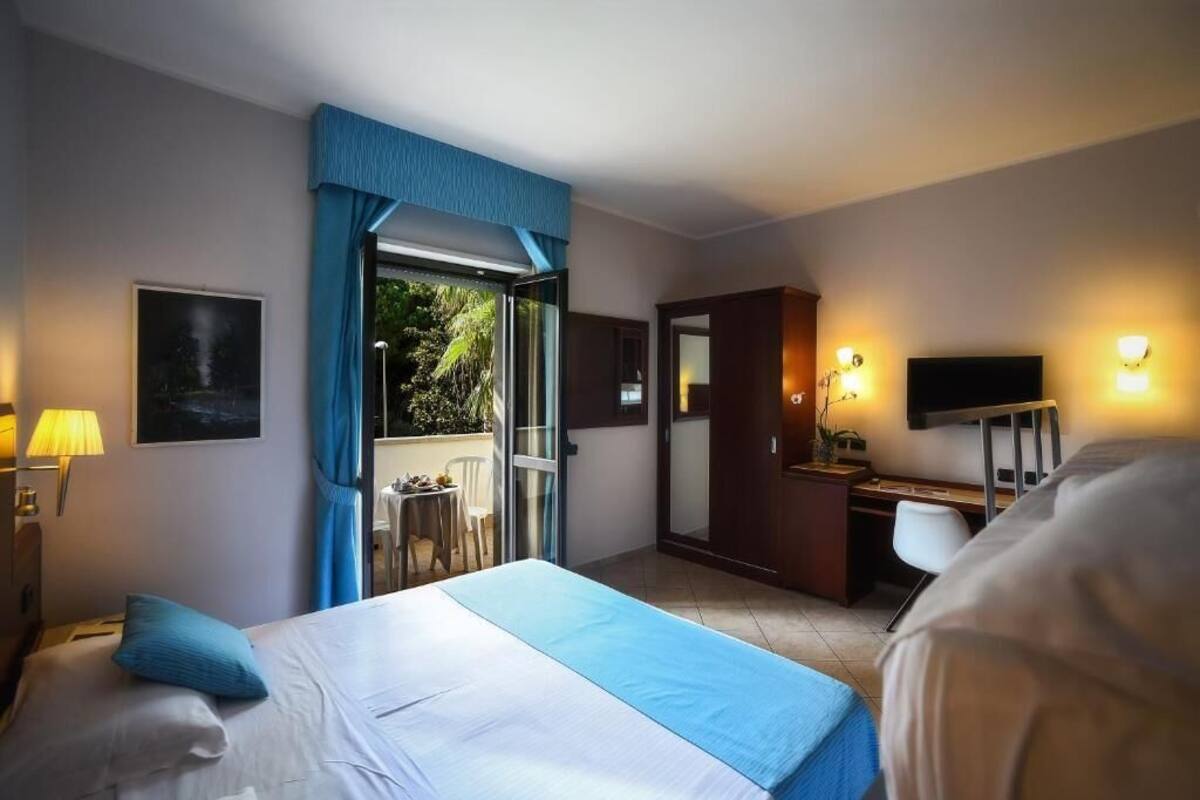 A hotel room with a bed, a desk, a chair, a television, and a balcony with a view of greenery.