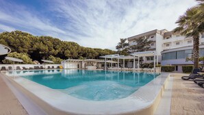 Una piscina con agua cristalina azul, rodeada de sillones de playa y sombrillas, con un edificio y árboles al fondo.