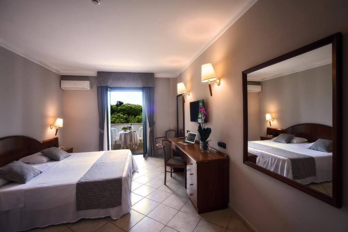 A hotel room with two beds, a desk, a chair, a TV, and a balcony with a view of greenery.