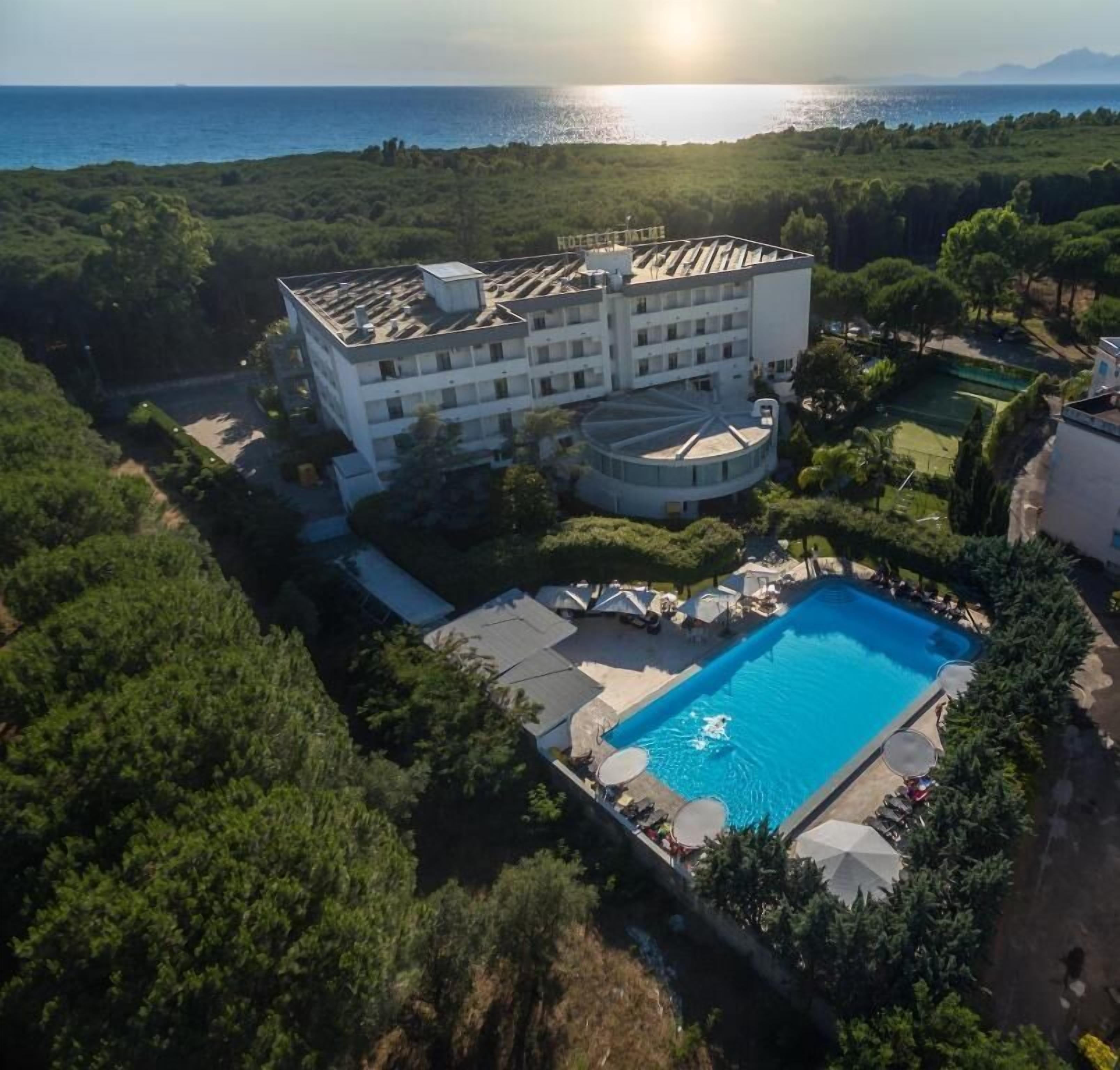 A hotel with a pool, surrounded by greenery and a view of the sea.