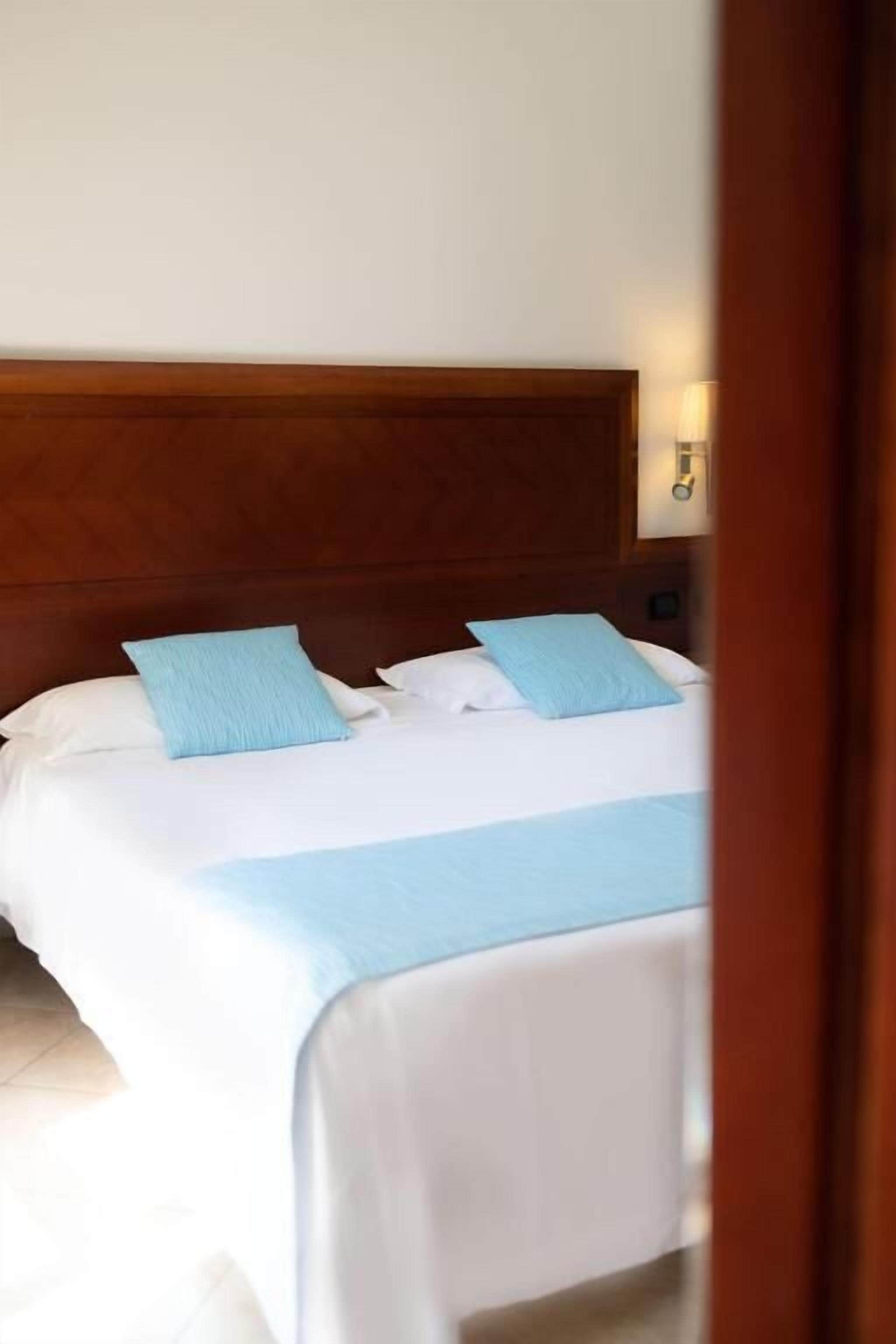 A neatly made bed with white linens and blue pillows, a wooden headboard, and a wall-mounted light fixture.