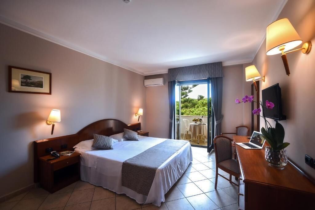 A hotel room with a bed, bedside lamps, a desk with a laptop, a chair, and a TV.