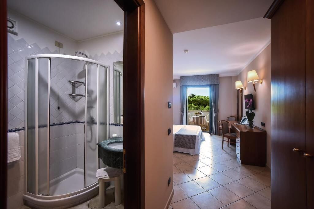 Un bagno d'hotel con doccia in vetro, lavandino e WC. Nell'altra stanza si trova un letto e una scrivania con una sedia.