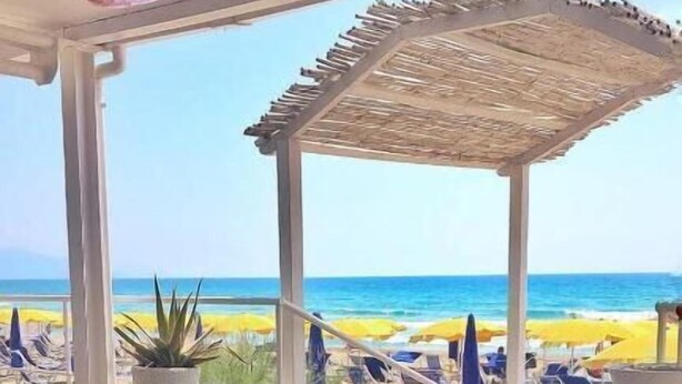 Un área de comedor junto a la playa con vista al océano, que incluye una pérgola y sillones de descanso.