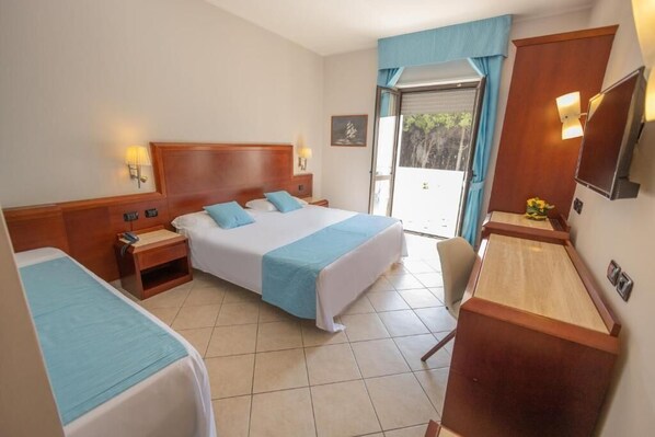 Une chambre d’hôtel avec un grand lit, un bureau et une chaise. Il y a une fenêtre avec des rideaux, et de la fenêtre, on peut admirer la verdure environnante.