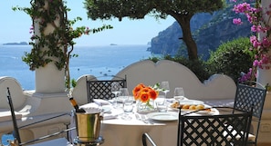 Mesa para dos con una botella de champán, copas y un plato de comida, con vistas a la costa, acantilados y el mar.