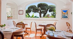 A dining area with a large window overlooking a garden, wooden furniture, and a framed painting on the wall.
