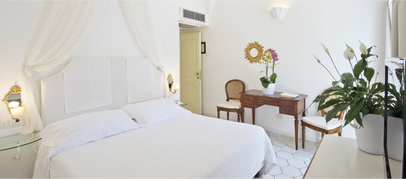 Une chambre avec un grand lit, un bureau et une chaise. On y trouve une plante en pot et un miroir fixé au mur.