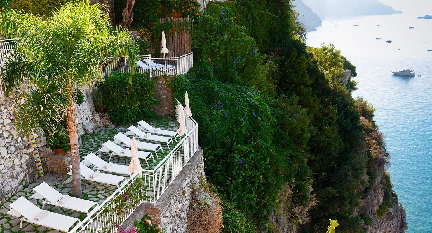 A cliffside terrace with white lounge chairs and umbrellas overlooking a coastal area with boats.