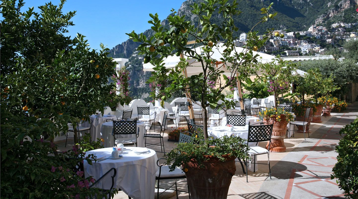 An outdoor dining area with round tables and chairs, surrounded by potted plants and trees.