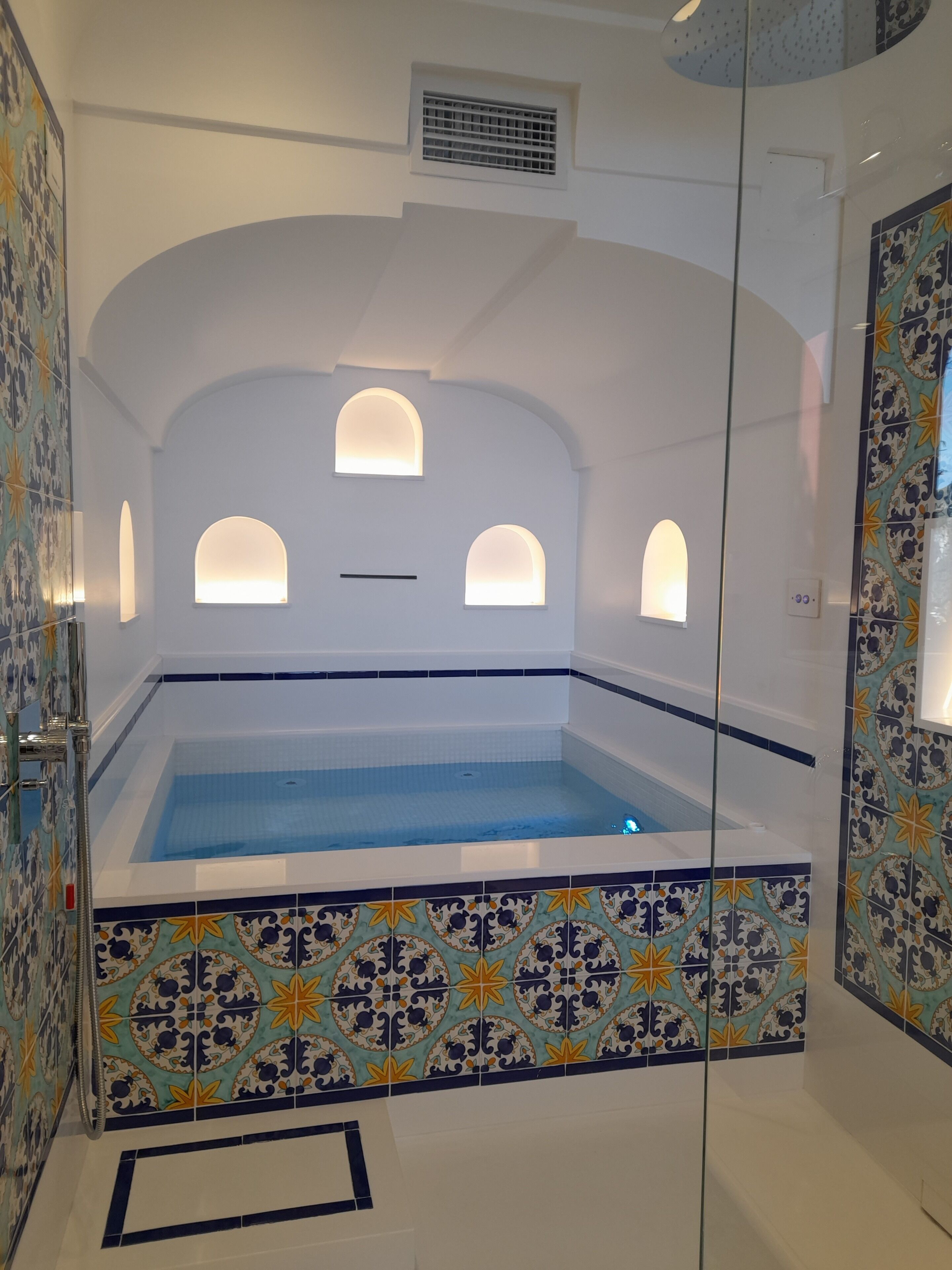 Una piscina interior moderna con cerramiento de vidrio transparente, paredes de azulejos decorativos e iluminación empotrada.