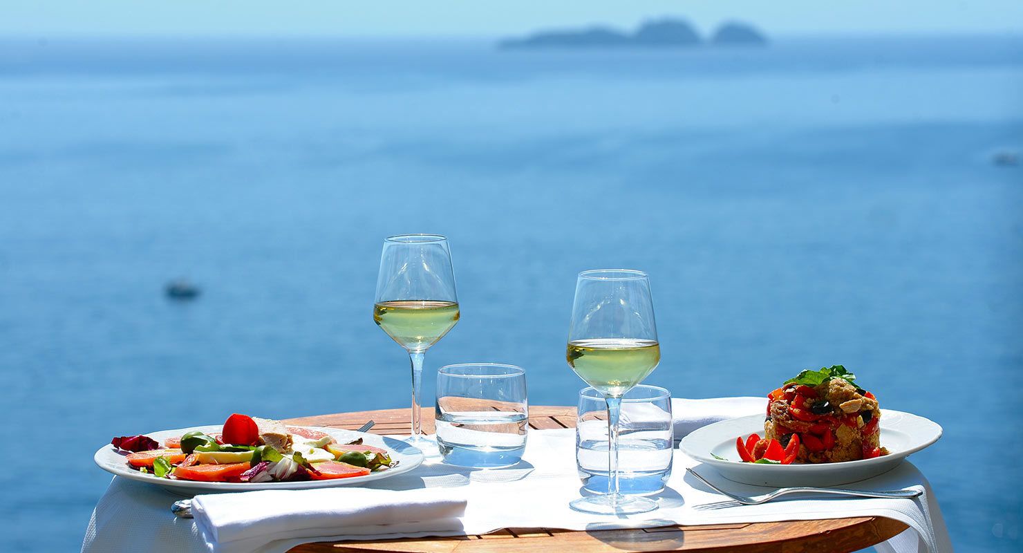 A table with two plates of food, two glasses of white wine, and a glass of water, set against a backdrop of a clear blue sea and distant islands.