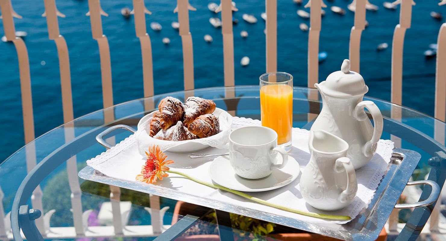 Bandeja de desayuno con pasteles, un vaso de jugo de naranja y tazas de té blancas junto con una jarrón sobre una mesa de vidrio con vista al mar.