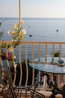 Un balcón con mesa y sillas de vidrio, con vista a un mar tranquilo y barcos.