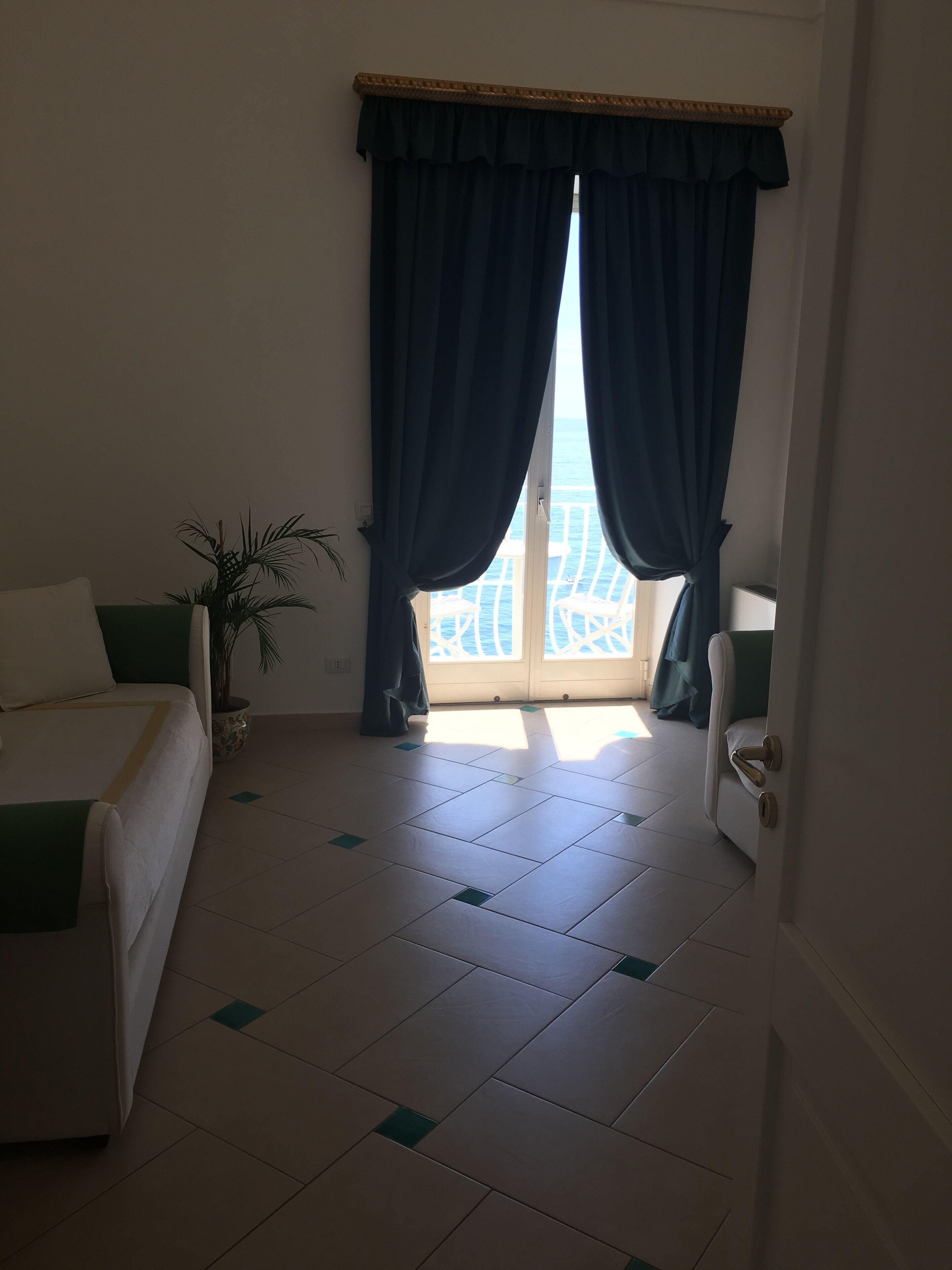 A bedroom with a bed, a potted plant, and a view of the sea through a window with curtains.