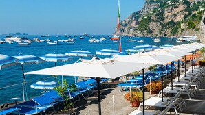 A coastal area with rows of sunbeds and umbrellas, boats on the water, and a rocky cliff in the background. - Marincanto (Positano)