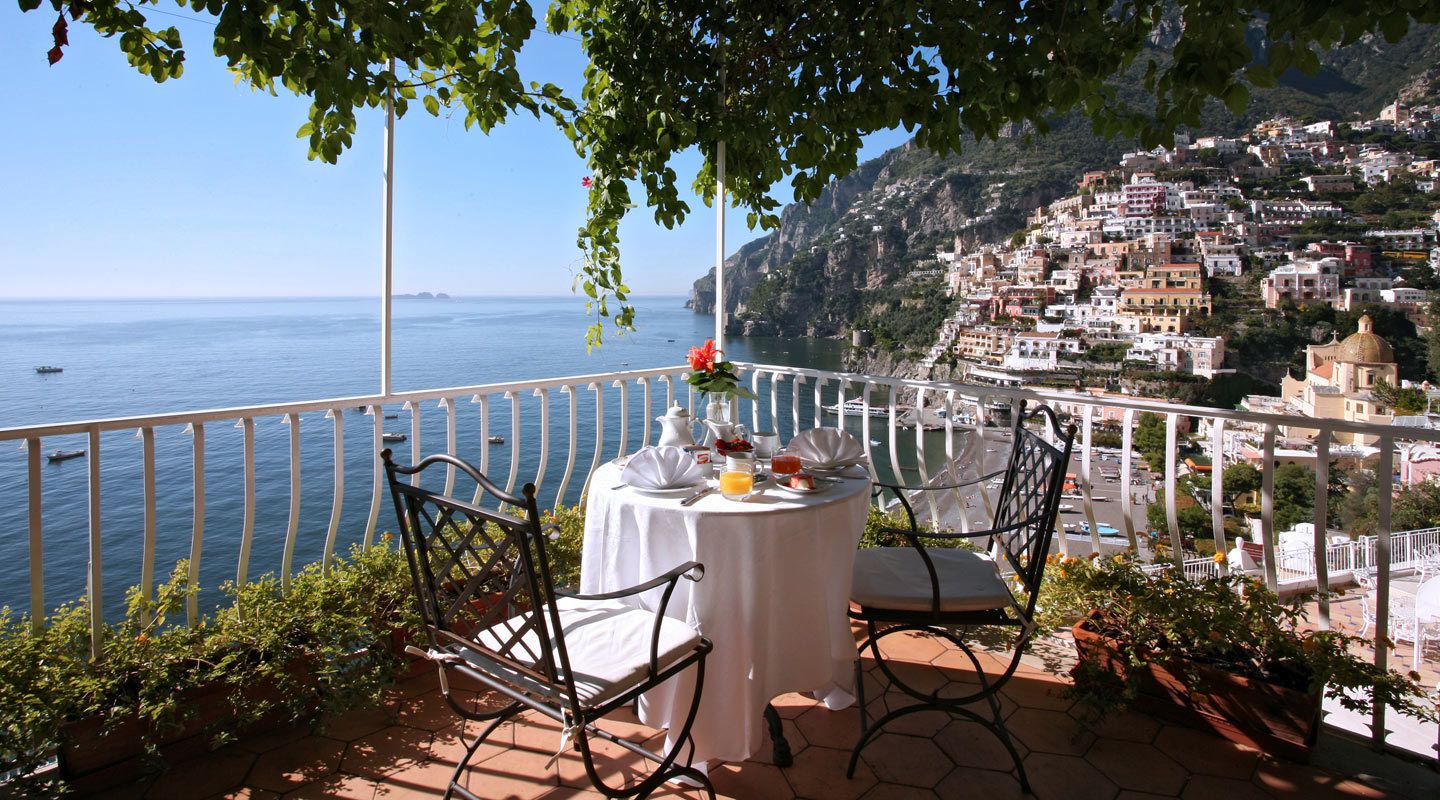 A terrace with a table set for two, overlooking a coastal town and the sea.