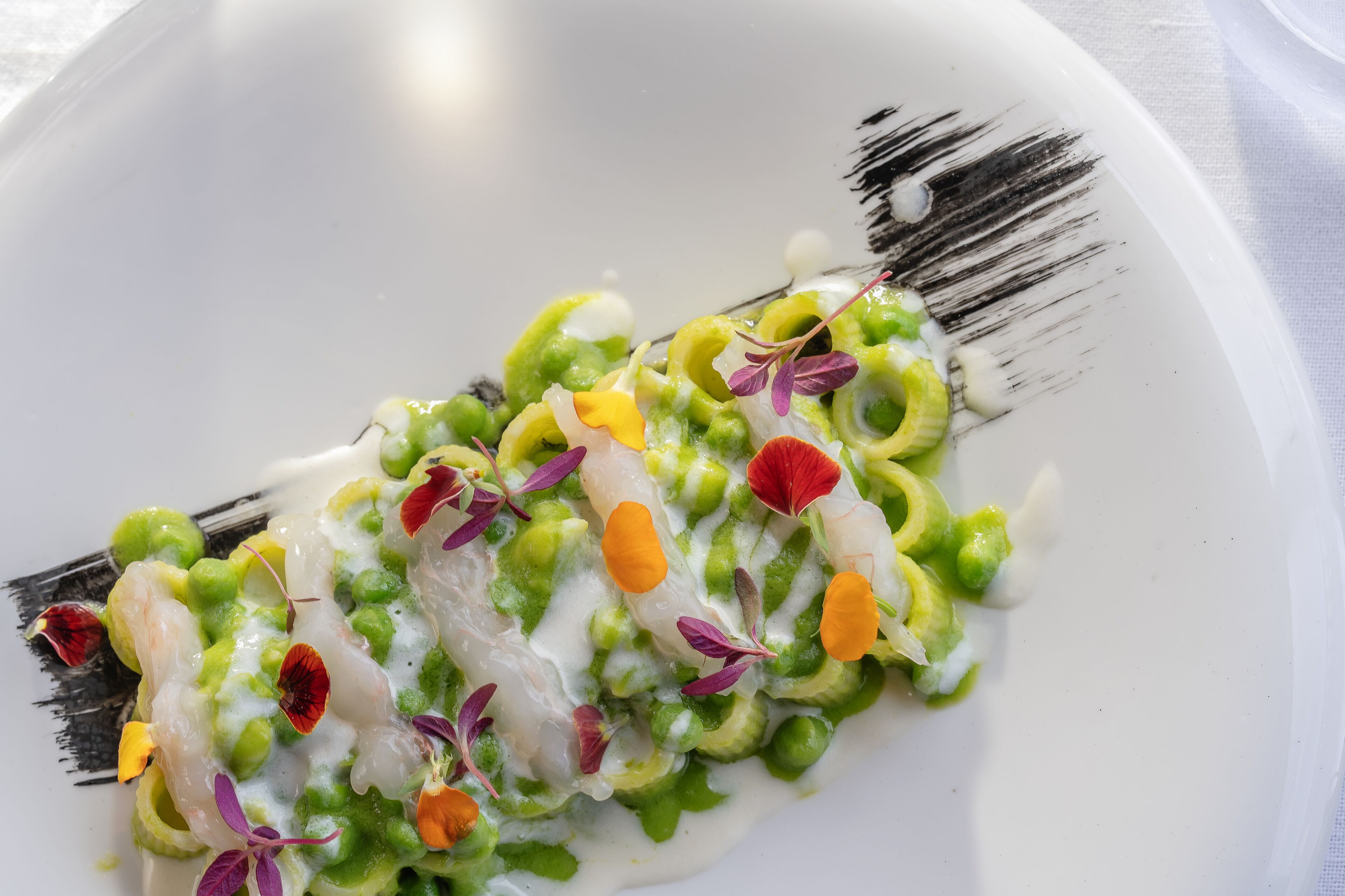 Um prato branco com um prato de frutos do mar, decorado com ervilhas verdes, camarões e flores comestíveis coloridas.