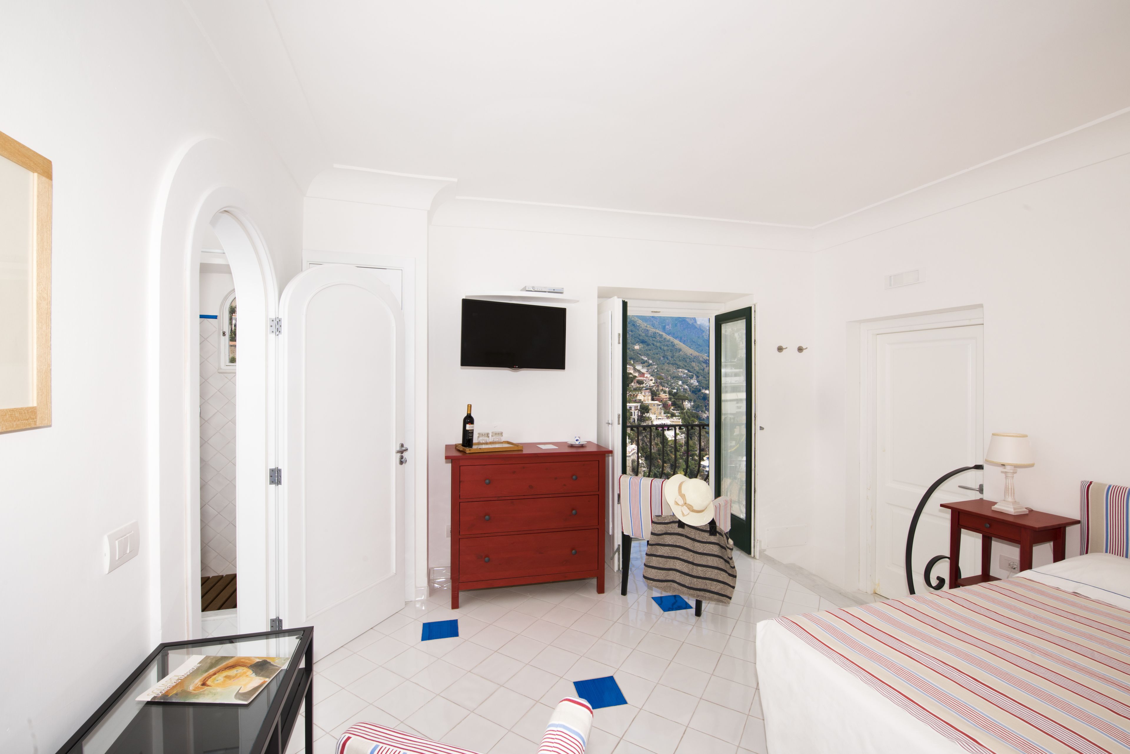 Quarto com cama, penteadeira, TV fixada na parede e vista para as montanhas por uma porta aberta.