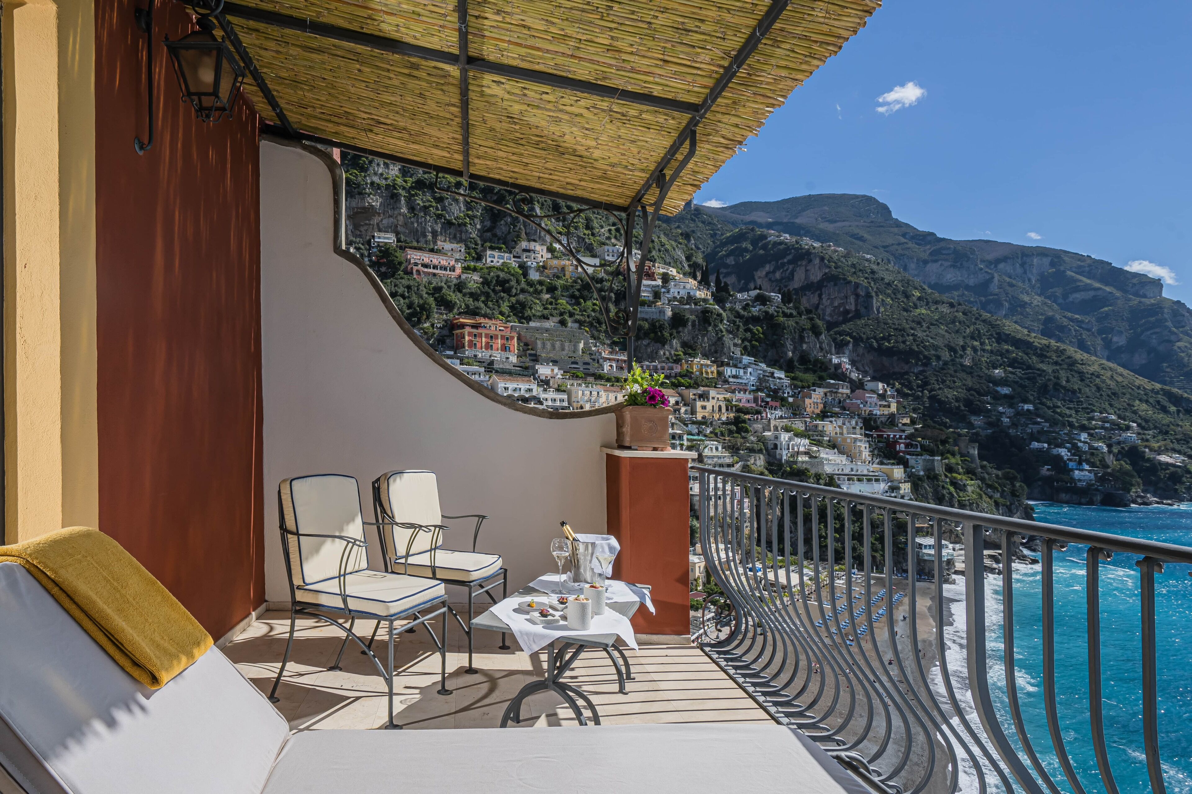 A balcony with a table set for dining, overlooking a coastal town with buildings on a hillside.